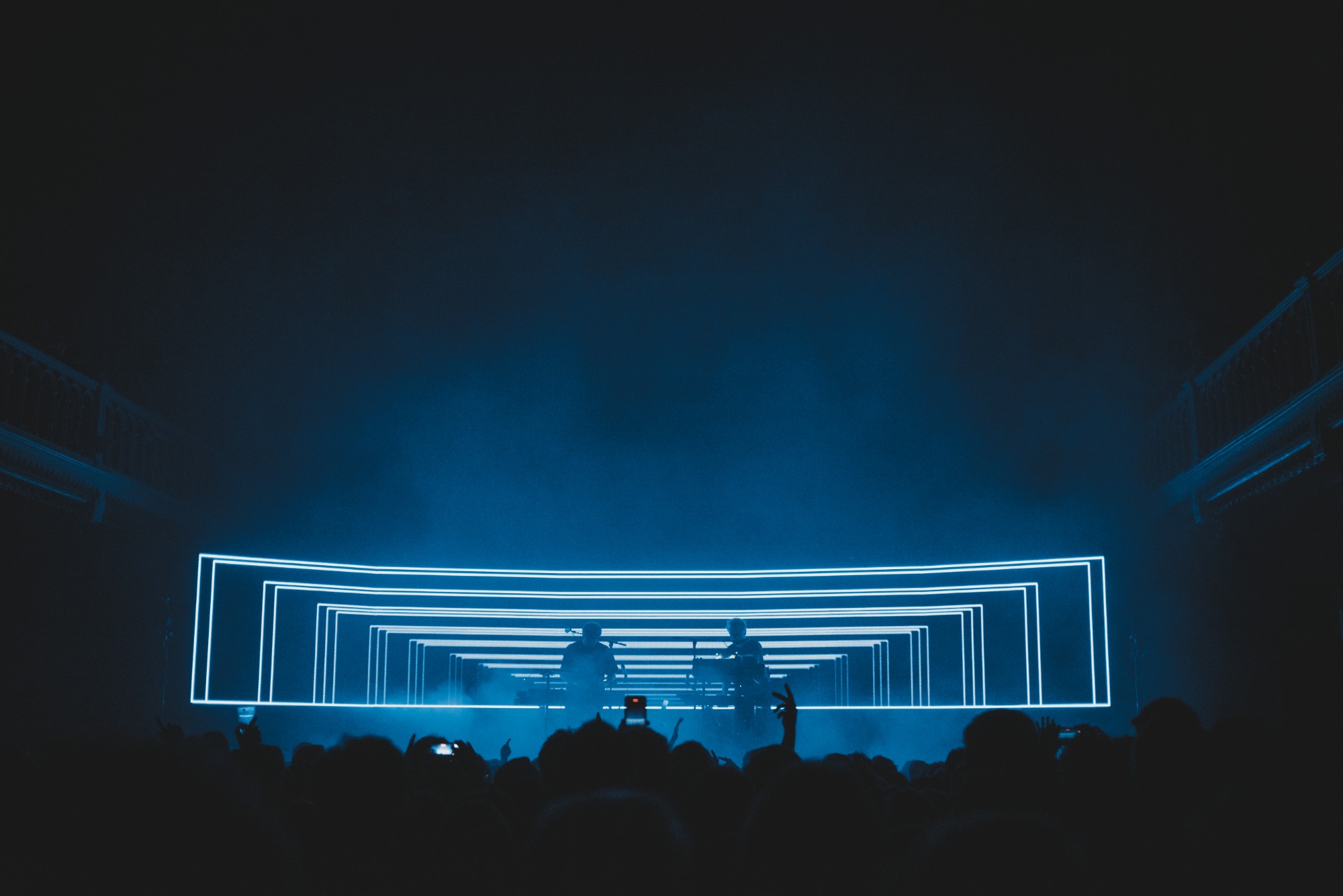 Een foto van achterin de zaal van Paradiso. We zien het publiek van achteren en de silhouetten van Weval. Ze worden van achter blauw belicht door een LED-scherm.