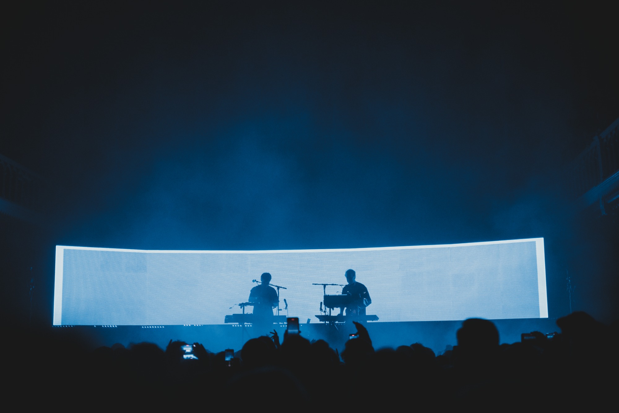 Een foto van achterin de zaal van Paradiso. We zien het publiek van achteren en de silhouetten van Weval. Ze worden van achter blauw belicht door een LED-scherm.