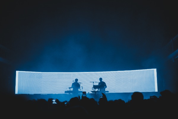 Een foto van achterin de zaal van Paradiso. We zien het publiek van achteren en de silhouetten van Weval. Ze worden van achter blauw belicht door een LED-scherm.