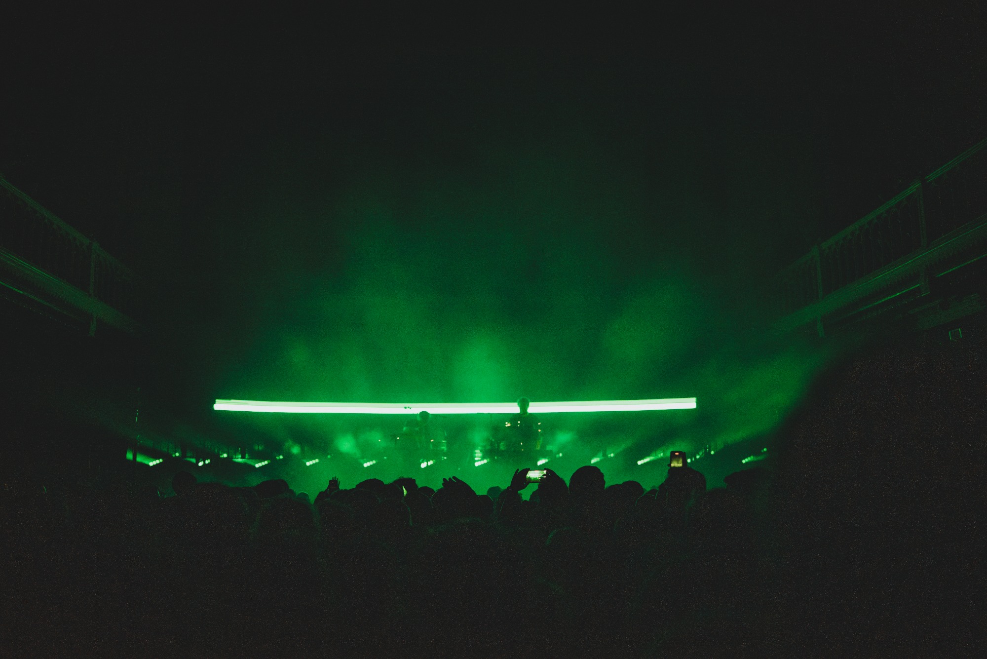 Een foto van achterin de zaal van Paradiso. We zien het publiek van achteren en de silhouetten van Weval. Ze worden van achter groen belicht door een streep op een LED-scherm.