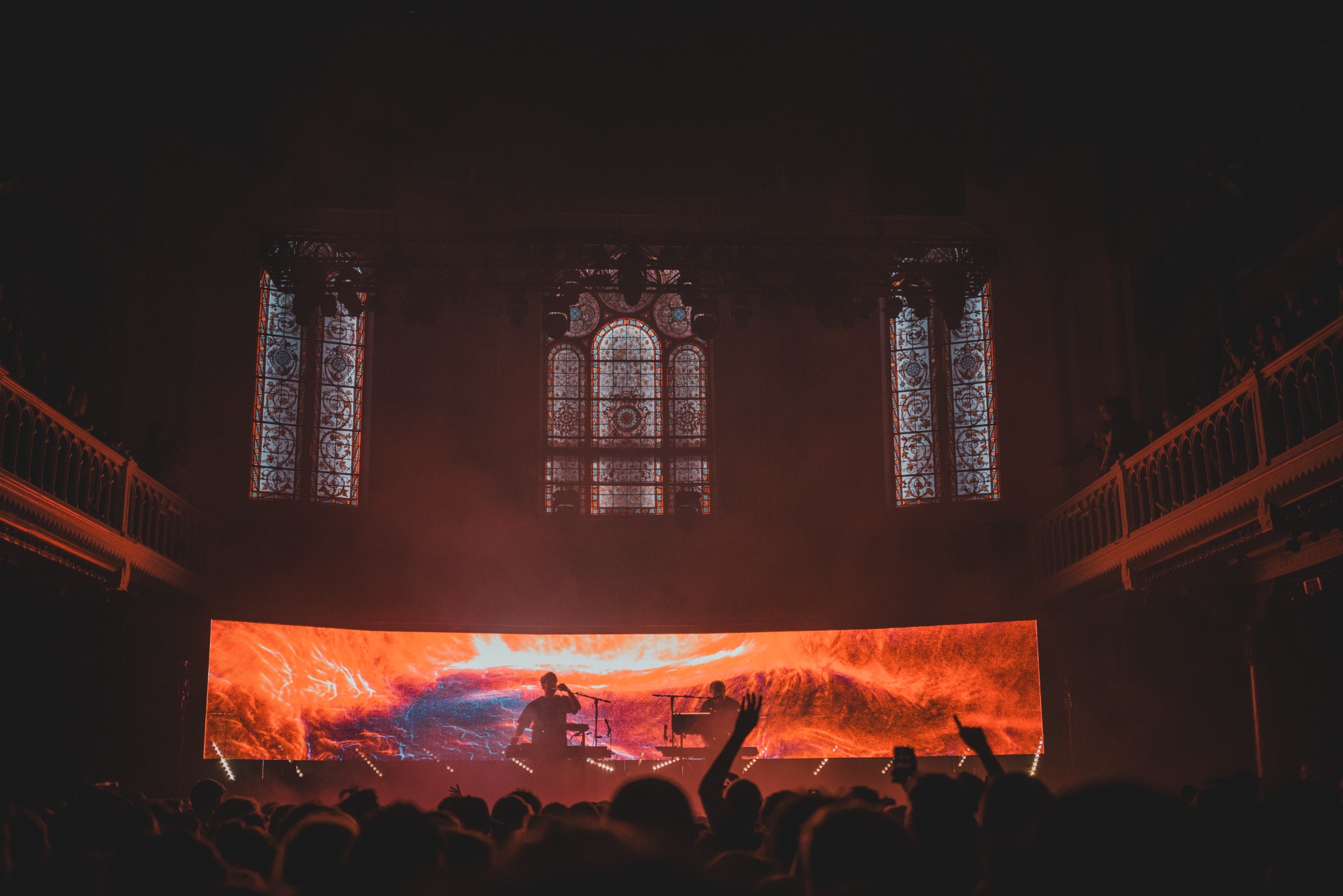 Een foto van achterin de zaal van Paradiso. We zien het publiek van achteren en de silhouetten van Weval. Ze worden van achter rood belicht door een visual op een LED-scherm.