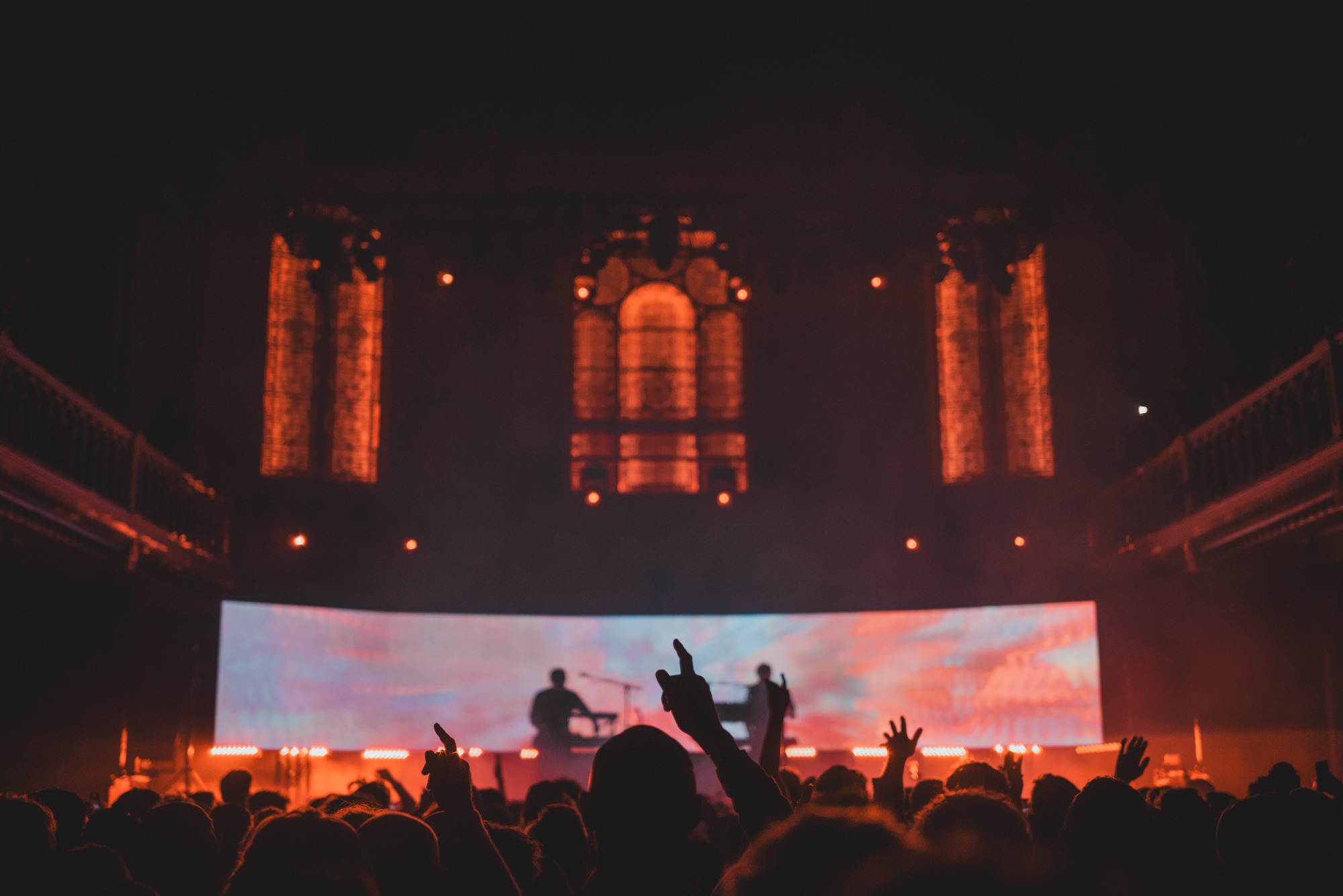 Een foto van achterin de zaal van Paradiso. We zien het publiek van achteren en de silhouetten van Weval. Ze worden van achter rood belicht door een visual op een LED-scherm.