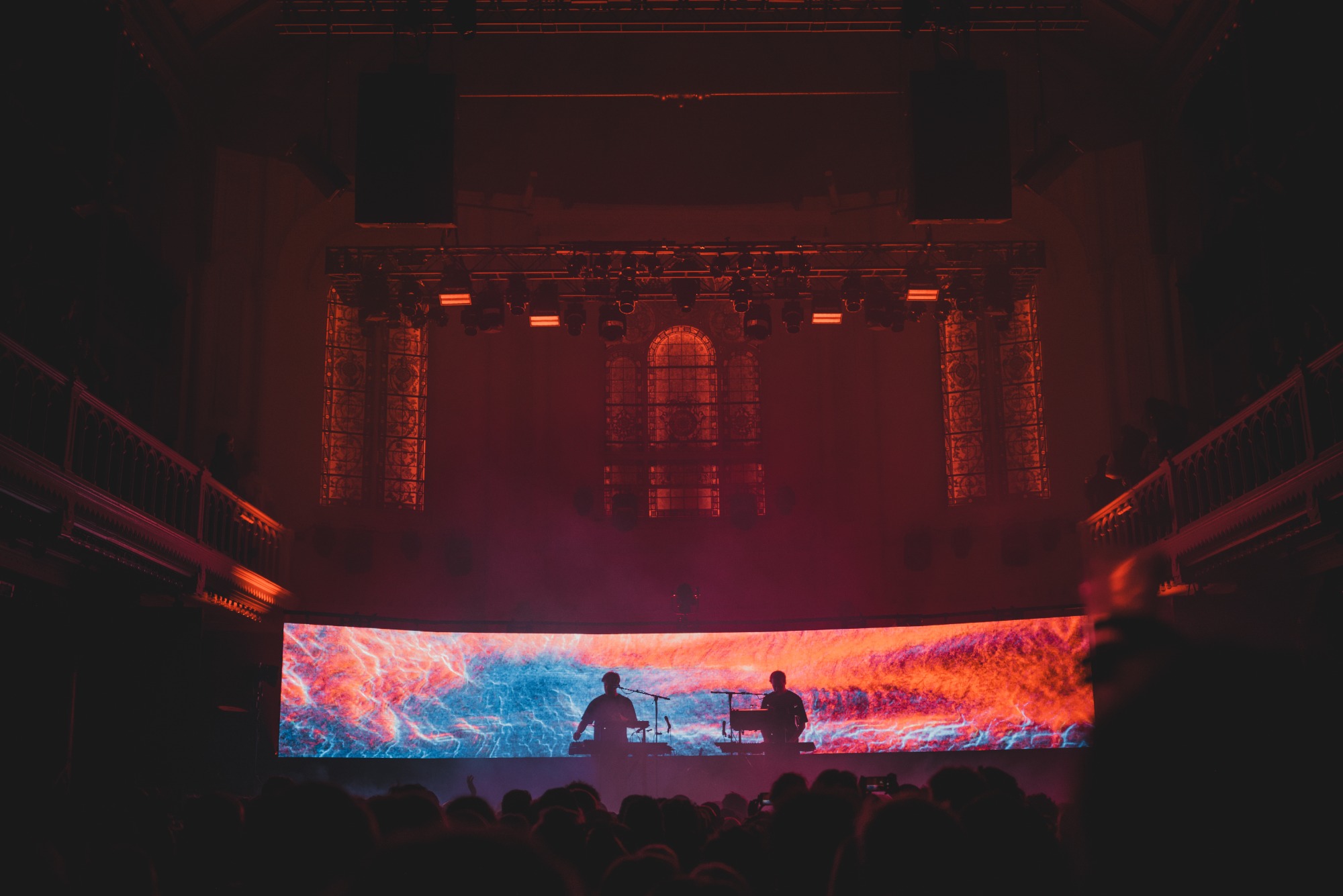 Een foto van achterin de zaal van Paradiso. We zien het publiek van achteren en de silhouetten van Weval. Ze worden van achter rood en blauw belicht door een visual op een LED-scherm.
