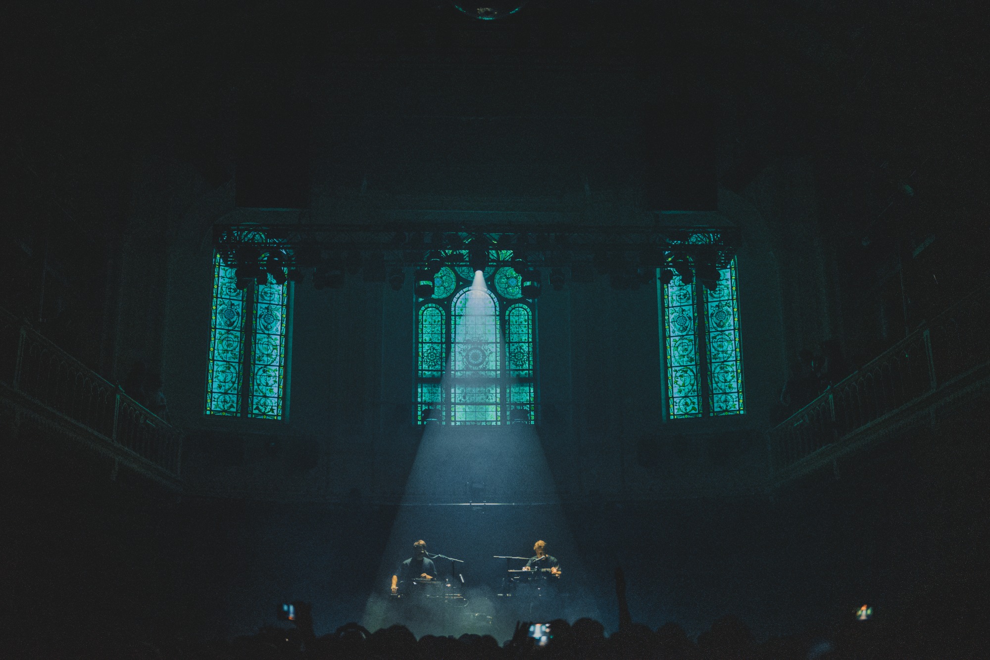 Een foto van achterin de zaal van Paradiso. Een spot richt van bovenin de zaal op het podium. We zien de kerkramen van Paradiso.
