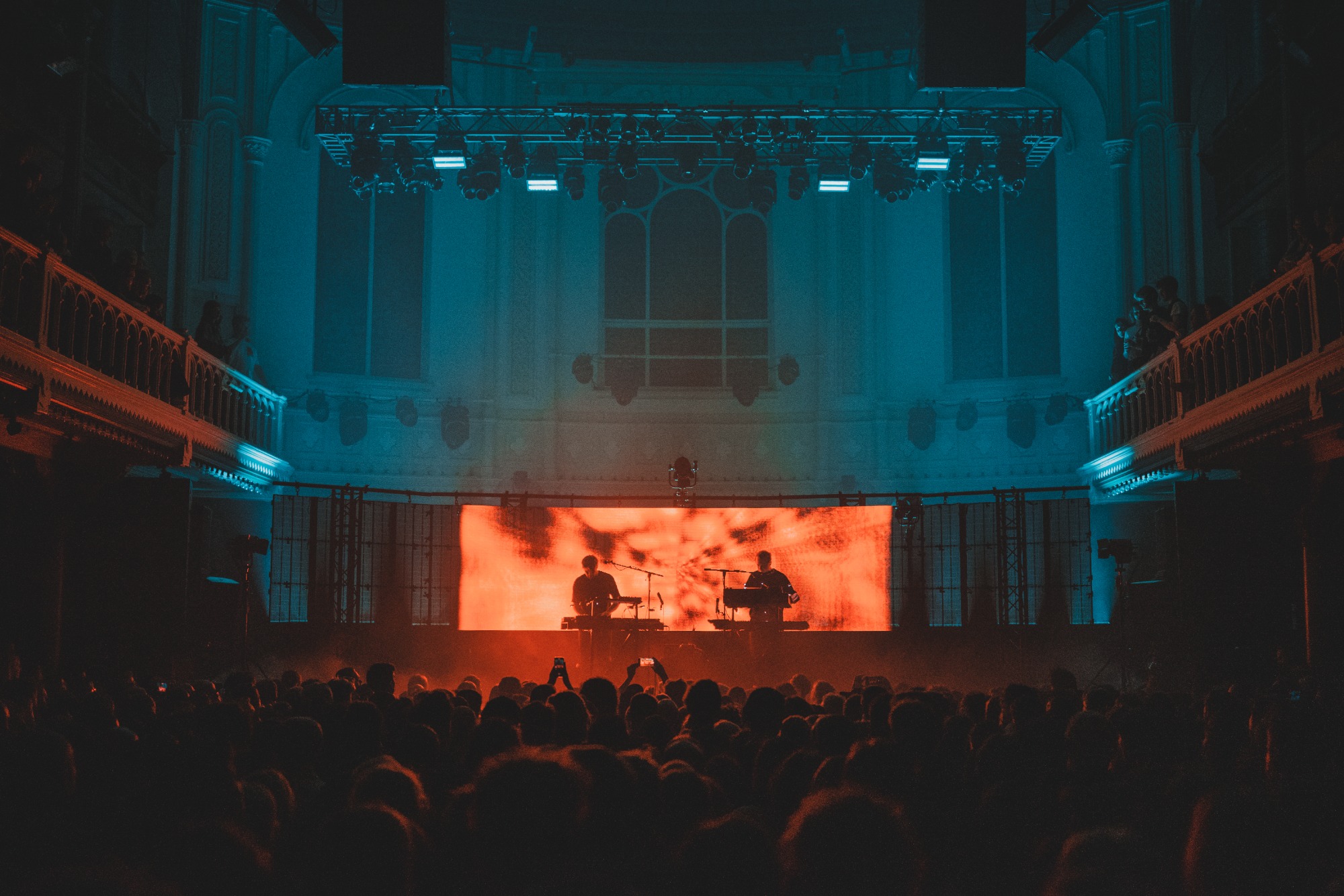 Een foto van achterin de zaal van Paradiso. We zien het publiek van achteren en de silhouetten van Weval. Ze worden van achter belicht door een LED-scherm.