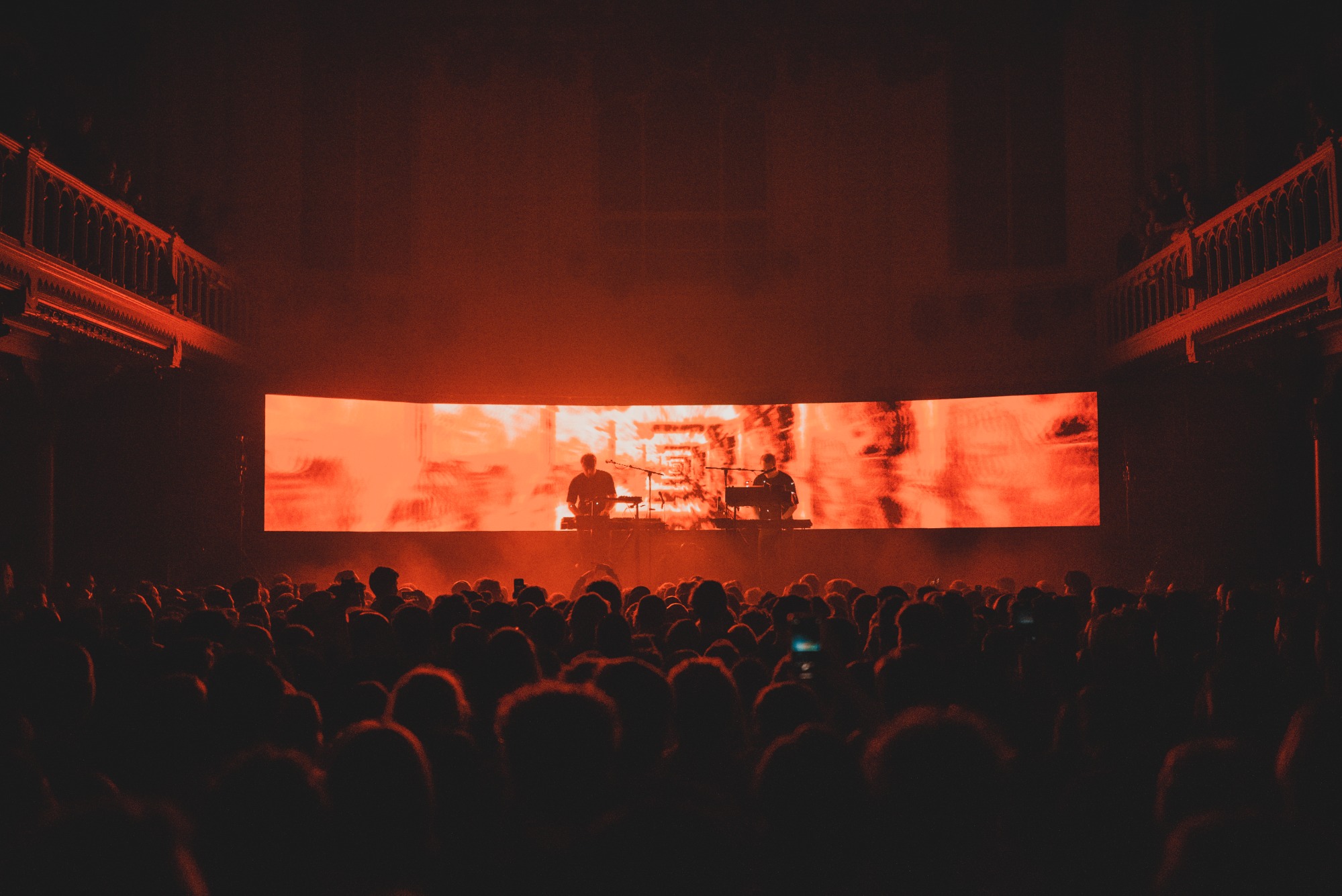Een foto van achterin de zaal van Paradiso. We zien het publiek van achteren en de silhouetten van Weval. Ze worden van achter belicht door een LED-scherm.