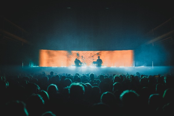 Een foto van achterin de zaal van Paradiso. We zien het publiek van achteren en de silhouetten van Weval. Ze worden van achter belicht door een LED-scherm.