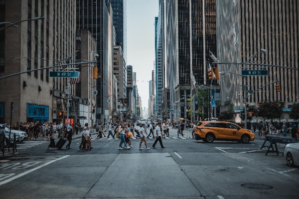 Een drukke straat in New York. We kijken de diepte in langs een aantal auto's en veel voetgangers